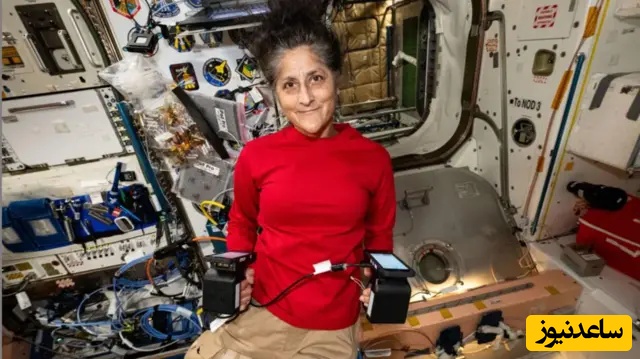 تصویر ویلیامز در داخل ایستگاه فضایی. او لباس سرخ بر تن دارد و در هر دو دستش تجهیزات را گرفته است.