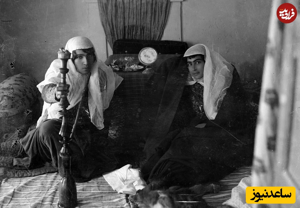 دو زن ایرانی عهد قاجار در کنار کرسی نشسته‌اند؛ یکی از آن‌ها قلیان می‌کشد و بر روی کرسی نیز   ظروف تنقلات به چشم می‌خورند